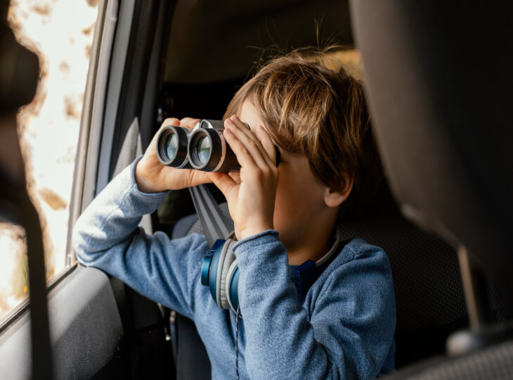 Petit garçon avec des écouteurs autour du cou observant le paysage avec des jumelles depuis l'intérieur d'un véhicule, à la recherche d'éléments pour cocher une des cases présentent sur son bingo voiture.
